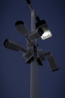 Several security cameras are mounted on a tall pole, with a bright light fixture attached nearby. The background is a dark evening sky.