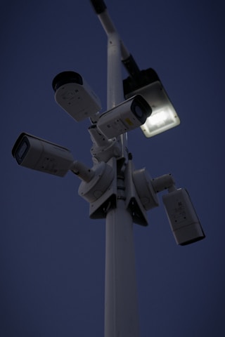 Several security cameras are mounted on a tall pole, with a bright light fixture attached nearby. The background is a dark evening sky.