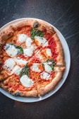 A round pizza topped with melted mozzarella cheese, fresh basil leaves, and tomato sauce, served on a white plate. The crust is well-baked with a golden-brown hue.