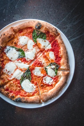 A round pizza topped with melted mozzarella cheese, fresh basil leaves, and tomato sauce, served on a white plate. The crust is well-baked with a golden-brown hue.