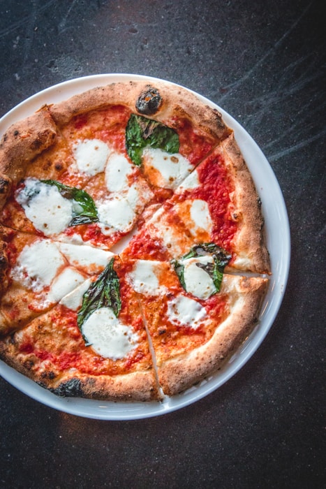 A round pizza topped with melted mozzarella cheese, fresh basil leaves, and tomato sauce, served on a white plate. The crust is well-baked with a golden-brown hue.