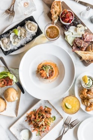 A table covered in a variety of gourmet dishes, including a plate of oysters on ice, a charcuterie board with cheeses and cured meats, a dish with a seafood topping, a plate with sliders, and a vibrant salad. A glass of beer and a cocktail with a lime wedge are also present, along with utensils and a small candle.