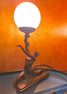 A decorative lamp with a bronze figurine of a woman in an elegant pose, kneeling with arms raised, holding a glowing spherical light. The background features a warm, textured orange surface.