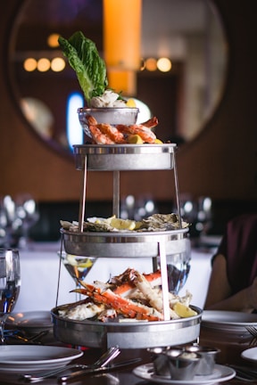 A three-tiered seafood platter features various shellfish including prawns, crab legs, and oysters, garnished with lemon wedges and leafy greens. This elegant presentation is set on a dining table with glassware and table settings, suggesting a fine dining experience.