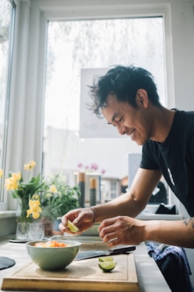 A person is preparing a meal in a bright kitchen, slicing a lime over a bowl of chopped vegetables on a wooden cutting board. Sunlight streams through a large window, illuminating yellow flowers in a vase nearby. The person appears focused and content while cooking.