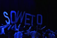 A group of people in dark clothing hold up large letters that spell 'SOWETO' against a dark background. The lighting is low, creating a dramatic and focused effect on the letters.