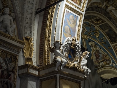 The image depicts an ornate interior with intricate architectural details, including sculpted cherubs and decorative gold accents. Painted panels with angelic faces complement the lavish design, featuring blue and gold hues in ornamental patterns.