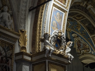 The image depicts an ornate interior with intricate architectural details, including sculpted cherubs and decorative gold accents. Painted panels with angelic faces complement the lavish design, featuring blue and gold hues in ornamental patterns.