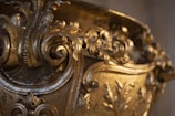 A close-up of an intricately designed golden object featuring detailed ornamental scrollwork and floral patterns. The craftsmanship is elaborate, with a shiny, polished gold finish and an ornate design with curving lines and embossed leaves.