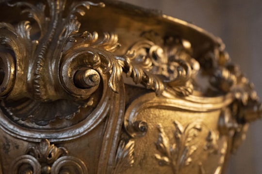A close-up of an intricately designed golden object featuring detailed ornamental scrollwork and floral patterns. The craftsmanship is elaborate, with a shiny, polished gold finish and an ornate design with curving lines and embossed leaves.