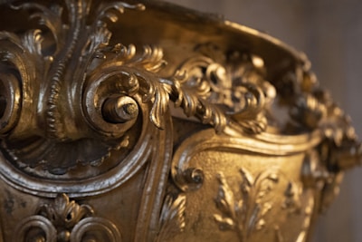 A close-up of an intricately designed golden object featuring detailed ornamental scrollwork and floral patterns. The craftsmanship is elaborate, with a shiny, polished gold finish and an ornate design with curving lines and embossed leaves.