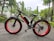 A black electric bicycle with large, wide tires and red wheel rims is parked on a paved path. The bike is labeled with 'VOLTEBYK' on the frame. It is surrounded by a lush, green park with trees and vegetation, and a backdrop of distant buildings and hills under an overcast sky.