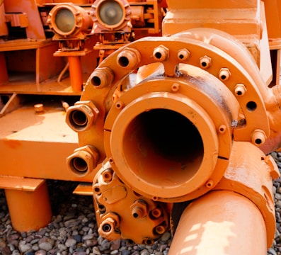 An industrial machine part painted in bright orange, featuring pipes, bolts, and connectors. The equipment is placed on a surface covered with small pebbles, with other mechanical components in the background.