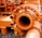 An industrial machine part painted in bright orange, featuring pipes, bolts, and connectors. The equipment is placed on a surface covered with small pebbles, with other mechanical components in the background.