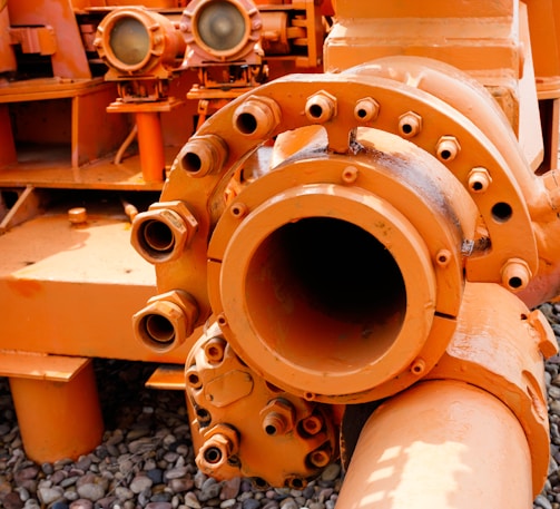 An industrial machine part painted in bright orange, featuring pipes, bolts, and connectors. The equipment is placed on a surface covered with small pebbles, with other mechanical components in the background.