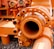 An industrial machine part painted in bright orange, featuring pipes, bolts, and connectors. The equipment is placed on a surface covered with small pebbles, with other mechanical components in the background.