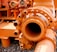 An industrial machine part painted in bright orange, featuring pipes, bolts, and connectors. The equipment is placed on a surface covered with small pebbles, with other mechanical components in the background.