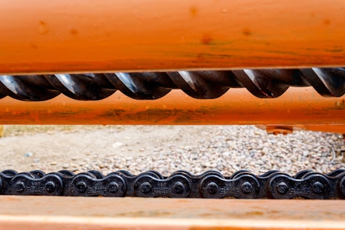 A close-up view of a mechanical structure featuring an orange metal surface with a prominent black spiral gear and a chain below it. The background is blurred, showing a gravelly surface.