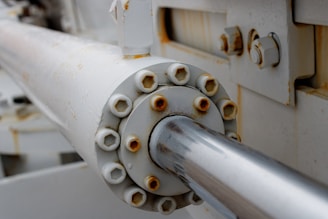 A close-up view of a metallic hydraulic or mechanical component with visible rust and bolts. The surface shows signs of wear and corrosion, indicating exposure to the elements. The structure is predominantly cylindrical with a shiny, reflective surface extending from the center.