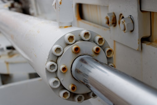 A close-up view of a metallic hydraulic or mechanical component with visible rust and bolts. The surface shows signs of wear and corrosion, indicating exposure to the elements. The structure is predominantly cylindrical with a shiny, reflective surface extending from the center.