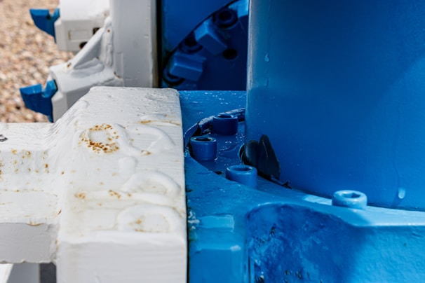 Close-up of a mechanical structure featuring interlocking white and blue components with visible rust spots and water droplets. The components appear to be part of a larger industrial machine.