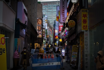 A bustling urban street scene with numerous illuminated signs featuring various languages, primarily Korean, advertising businesses such as restaurants and bars. Pedestrians wearing casual clothing are walking through the narrow pathway, bordered by temporary blue fencing and traffic cones. The atmosphere is lively, suggestive of a popular nightlife area.