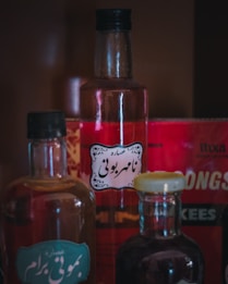 A collection of glass bottles filled with various colored liquids. Labels with intricate designs are attached to the bottles, and they are set against a deep red background. The image depicts a vintage or apothecary-like aesthetic with a focus on the bottles&rsquo; shapes and colors.