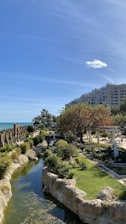 A landscaped park area featuring a lush garden with a variety of trees and shrubs, a small waterway bordered by rocks, and a clear blue sky overhead. In the background, there is a modern apartment building and a glimpse of the ocean. The environment is serene and picturesque, with well-maintained greenery and architectural elements like a bridge and columns.