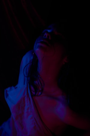 A person partially clothed in a loose shirt is illuminated by dramatic, colored lighting, with shadows obscuring parts of their face and upper body. The lighting is primarily in hues of blue and pink, creating a dramatic and possibly emotional atmosphere. The person's head is tilted back, with hair hanging down.