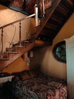 A cozy interior space features a rustic wooden staircase with wrought iron railings. Beneath the staircase, a small bed with a patterned quilt is tucked into the corner, accompanied by pillows. The walls are painted in warm tones with a decorative mirror hanging nearby. Soft lighting from wall-mounted lamps creates a warm ambiance.