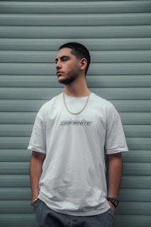A man stands against a background of horizontal teal panels, wearing a white t-shirt with 'Off-White' lettering, a gold chain, and a wristwatch. His hands are in his pockets, and he looks to the side.