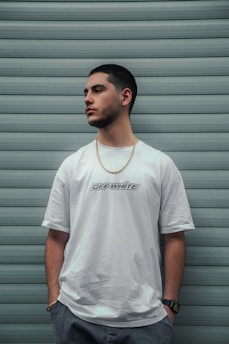 A man stands against a background of horizontal teal panels, wearing a white t-shirt with 'Off-White' lettering, a gold chain, and a wristwatch. His hands are in his pockets, and he looks to the side.