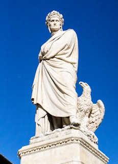 A tall marble statue of a robed figure wearing a laurel wreath stands prominently against a clear blue sky. Next to the figure is an eagle resting on the pedestal.