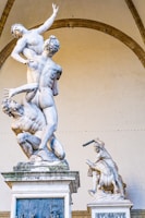 Two sculptures are prominently displayed, with one showing dynamic human figures in a dramatic, intertwined pose, while the other features a warrior-like figure with a shield and weapon. The statues are intricately detailed, capturing muscular forms and expressions, and are set against an arched backdrop.
