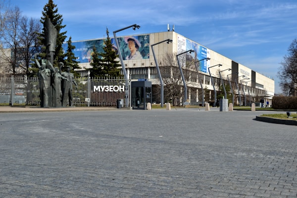 A large complex with a modern design featuring tall streetlights and a paved area in the foreground. A prominent statue made of dark material stands near the entrance, surrounded by trees and a fence. Large banners are displayed on the building's facade, and the word 'МУЗЕОН' is visible. The sky is clear with scattered clouds.