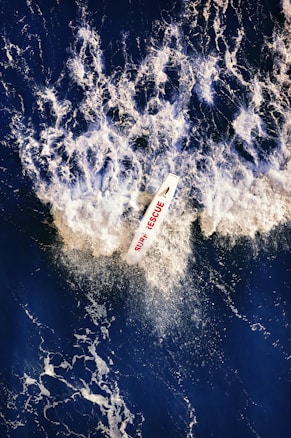 Aerial view of a surf rescue boat cutting through dark blue ocean water, creating a dramatic splash of white foam around it. The text 'SURF RESCUE' is visible on the boat in red letters.
