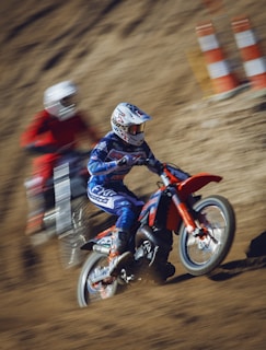 Two motocross riders are racing on a dirt track. The rider in the foreground is dressed in blue gear with a matching helmet and is riding a red dirt bike. The second rider in the background wears red gear and appears slightly blurred, emphasizing motion. Orange and white traffic cones are seen in the background.