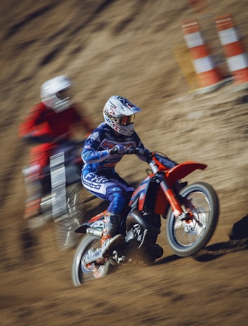 Two motocross riders are racing on a dirt track. The rider in the foreground is dressed in blue gear with a matching helmet and is riding a red dirt bike. The second rider in the background wears red gear and appears slightly blurred, emphasizing motion. Orange and white traffic cones are seen in the background.