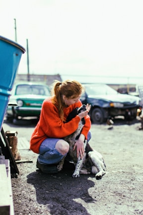 A person with red hair and a bright orange sweater crouches down to affectionately engage with a black and white dog. The setting appears to be outdoors with a few cars and a clear, bright sky in the background. The person is wearing ripped jeans and the interaction conveys a sense of warmth and companionship.