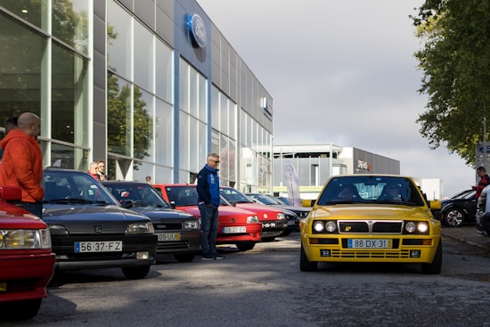 Several cars are parked in a lot near a building with a Ford logo. A notable yellow car is prominently driving toward the foreground. People are standing nearby, observing and interacting. The setting suggests a car meet or gathering, with a mix of different car models and colors.