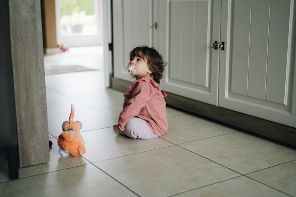 A toddler with a pacifier sits on a tiled floor near a cabinet, looking upwards. The child is wearing a pink top and white pants, and there is an orange plush toy rabbit nearby. The setting appears to be a kitchen or a hallway with natural light coming from an adjacent room.