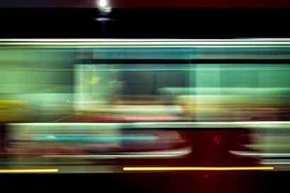 A blurred motion capture of a moving subway or train, characterized by streaks of light and color that create a sense of speed and dynamism. The image features a blend of green, yellow, and red hues with black background elements, conveying the fast-paced urban environment.