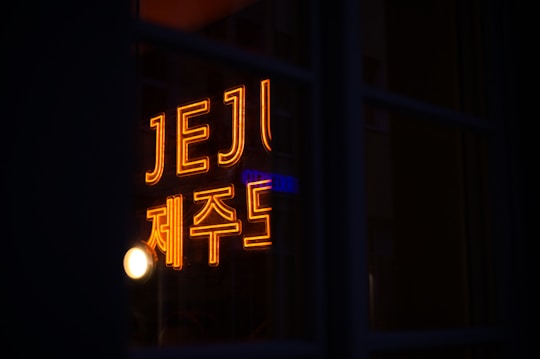 A bright neon sign with the word JEJU and Korean characters glows in a warm orange color, partially reflected in the window with a dark background.