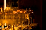A series of elegant wine glasses reflect warm ambient light, creating a shimmering effect. The background is softly blurred, highlighting the glasses in sharp focus.