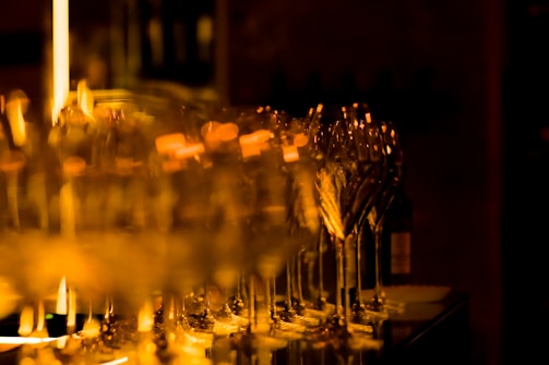 A series of elegant wine glasses reflect warm ambient light, creating a shimmering effect. The background is softly blurred, highlighting the glasses in sharp focus.
