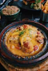 A delicious-looking dish served in a rustic ceramic pot, featuring tender chicken pieces in a rich, yellow curry sauce garnished with herbs and accompanied by vibrant sliced peppers and cherry tomatoes. Surrounding the main dish are additional plates with couscous and crispy French fries.