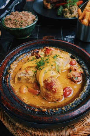 A delicious-looking dish served in a rustic ceramic pot, featuring tender chicken pieces in a rich, yellow curry sauce garnished with herbs and accompanied by vibrant sliced peppers and cherry tomatoes. Surrounding the main dish are additional plates with couscous and crispy French fries.