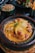 A delicious-looking dish served in a rustic ceramic pot, featuring tender chicken pieces in a rich, yellow curry sauce garnished with herbs and accompanied by vibrant sliced peppers and cherry tomatoes. Surrounding the main dish are additional plates with couscous and crispy French fries.