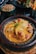 A delicious-looking dish served in a rustic ceramic pot, featuring tender chicken pieces in a rich, yellow curry sauce garnished with herbs and accompanied by vibrant sliced peppers and cherry tomatoes. Surrounding the main dish are additional plates with couscous and crispy French fries.