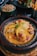 A delicious-looking dish served in a rustic ceramic pot, featuring tender chicken pieces in a rich, yellow curry sauce garnished with herbs and accompanied by vibrant sliced peppers and cherry tomatoes. Surrounding the main dish are additional plates with couscous and crispy French fries.